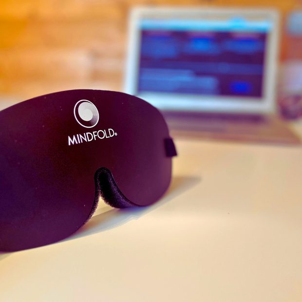 Antifaz Mindfold apoyado sobre una mesa, con un portátil desenfocado al fondo